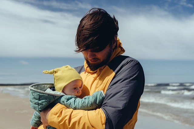 Pappa med spädbarn i famnen, på en strand.