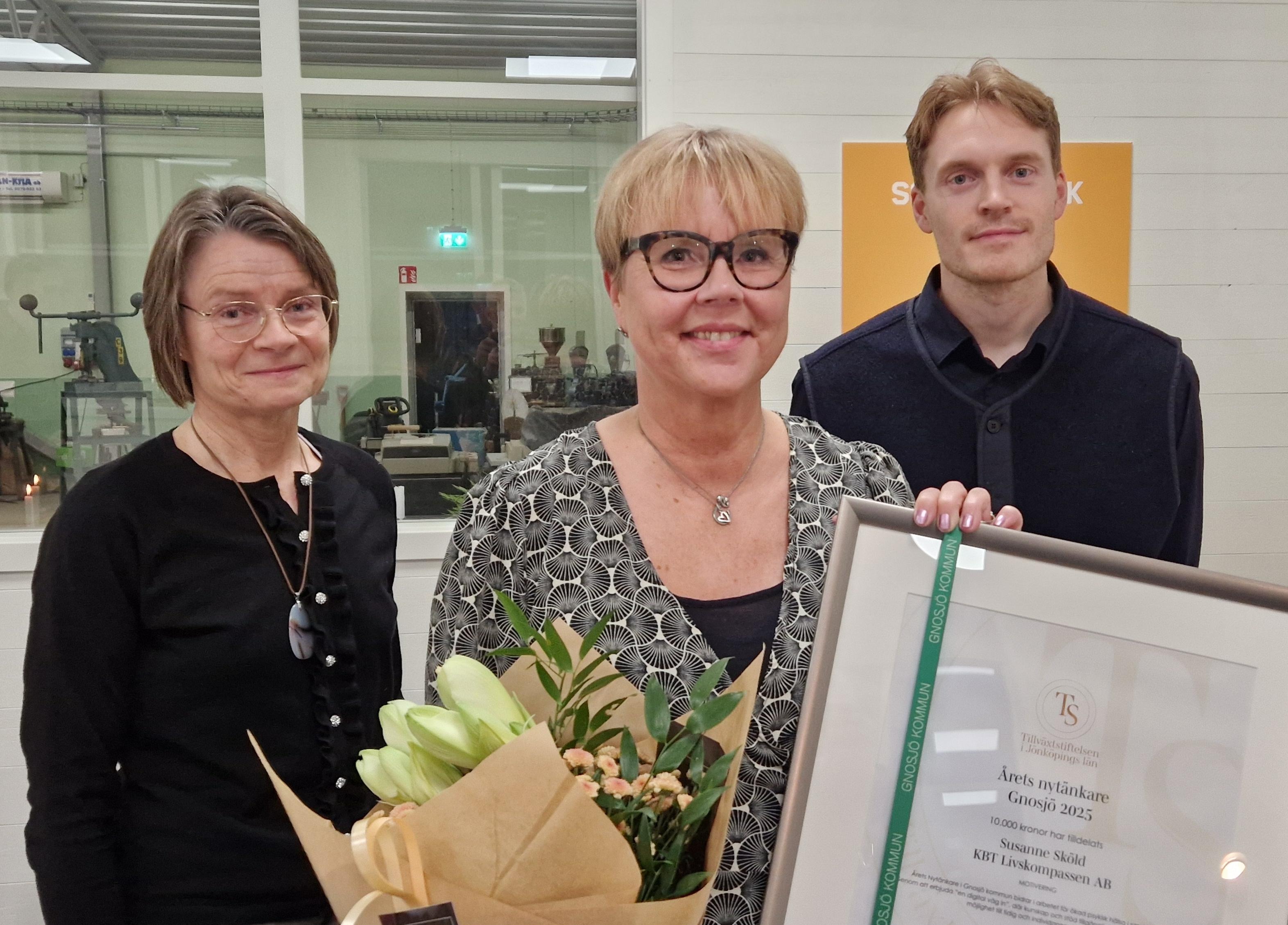 En kvinna som håller i ett inramat diplom och en blomma, vid sidan om henne står en man och en kvinna.