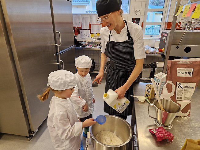 En kvinna  och två barn i kockkläder häller ingredienser i en bunke för att baka lunsar.