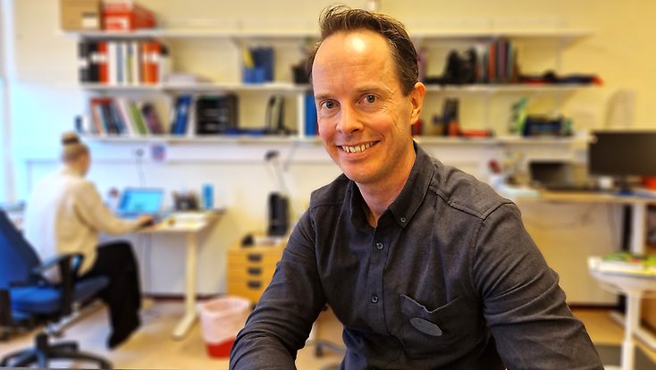 En glad man sitter i ett personalrum där en annan lärare sitter och förbereder en lektion.