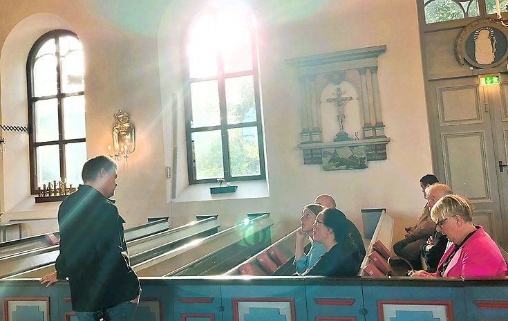 En man står i mittgången i kyrkan och pratar med personer i kyrkbänkarna.