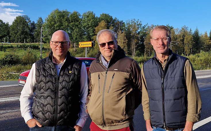 Tre män som står på en busshållplats vid en landsväg.