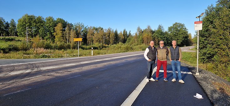 Tre män står på en busshållplats vid en landsväg.
