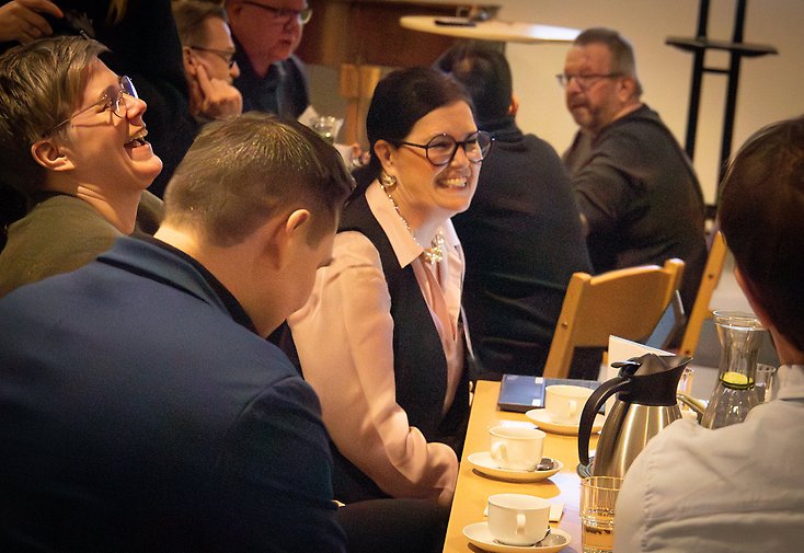 Kvinna som ler, runt om ett bord med kaffe och kaffekoppar sitter ett flertal personer.