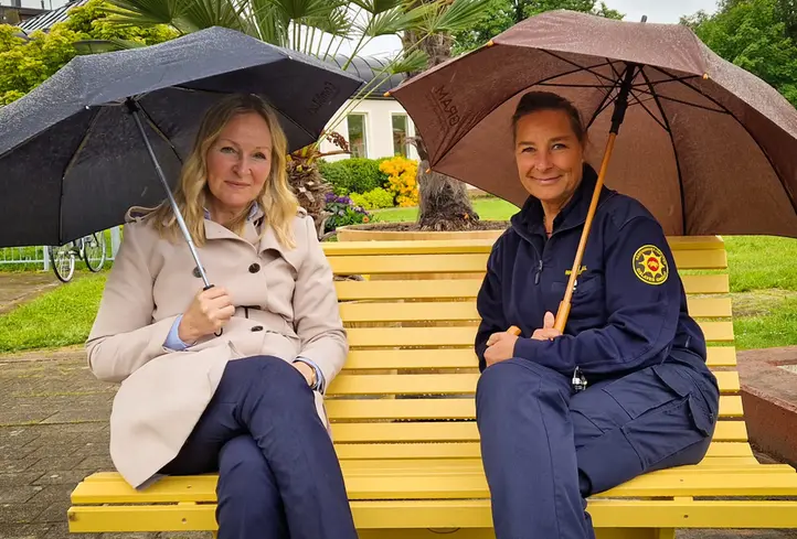 Två kvinnor med paraplyer som sitter på en gul parkbänk.