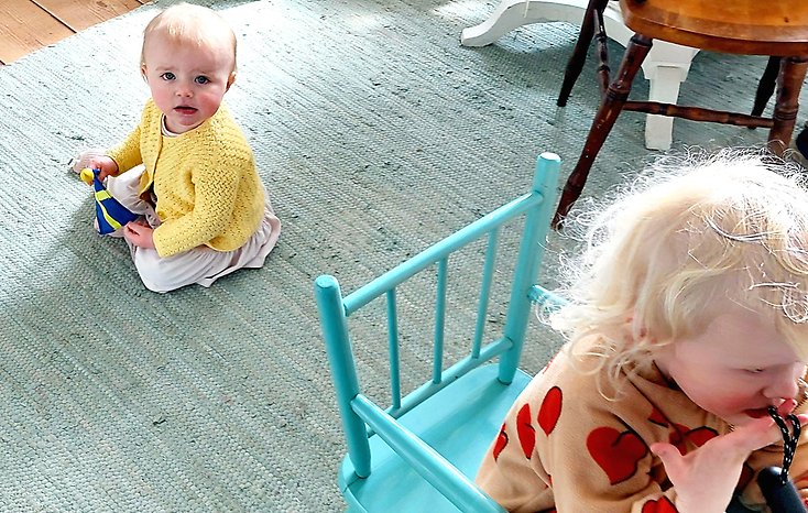 En liten baby sitter på golvet, framför sitter storasyster i en barnstol och vänder ryggen till lillasyster.